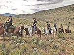 A line of horses and riders can be seen on a dusty mountain trail.